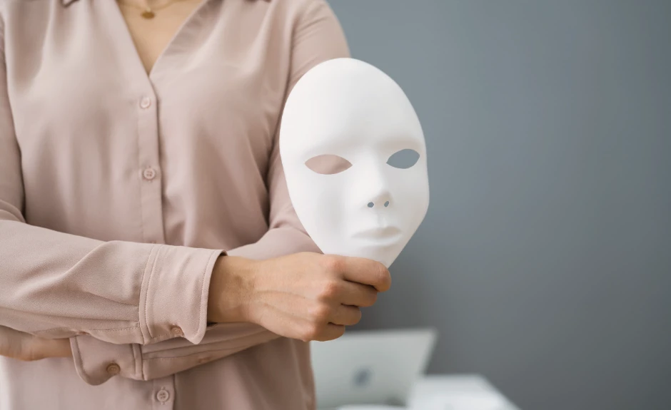 EIne Frau hält mit verschränkten Armen eine weiße Maske eines Gesichts in der Hand.