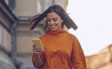 Frau mit orangem Pullover schaut im gehen auf ihr Handy und lächelt.