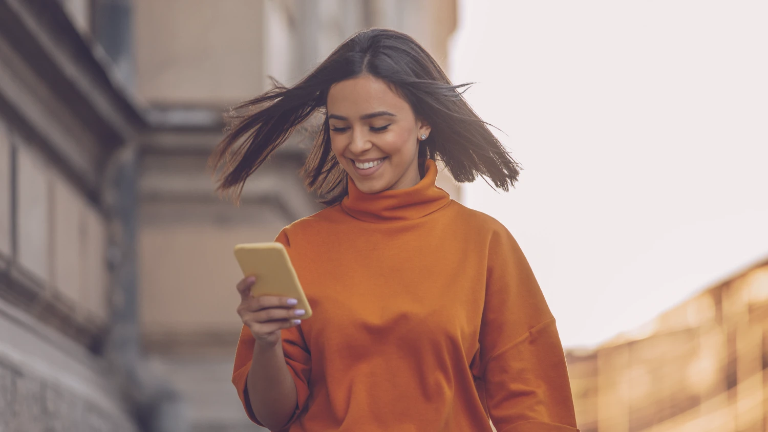 Frau mit orangem Pullover schaut im gehen auf ihr Handy und lächelt.