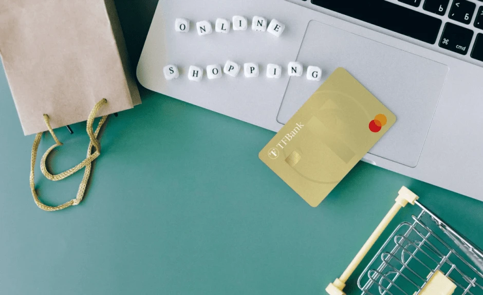 TF Mastercard Gold liegt auf Laptop. Daneben liegen kleine Würfel mit Buchstaben zusammengesetzt als "Online Shopping". Außerdem liegt dort eine kleine Tüte und eiin kleiner modelhafter Einkaufkorb.