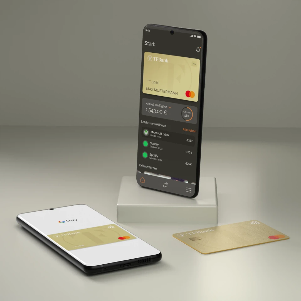 Smartphone steht in einem Raum auf einem Sockel. Es wird der Home Bildschirm der TF Bank Mobile App angezeigt. Neben dem Sockel liegt ein weiteres Smartphone mit der Google Pay Funktion, sowie eine physische TF Mastercard Gold.