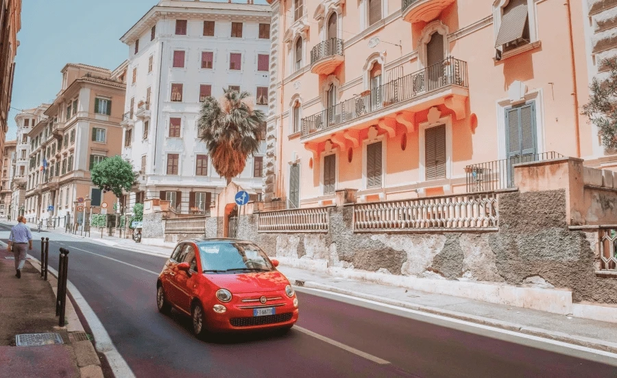 Auf einer Straße fährt ein rotes Auto. Im Hintergrund sind Häuser im südländischem Stil zu sehen.