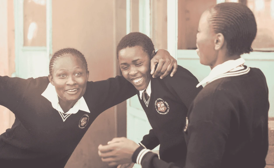 Es sind drei afrikanische Schüler zu sehen. Sie lachen und eine Person stützt sich auf den Schultern einer anderen.