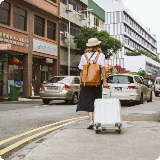 Eine Frau läuft auf dem Gehweg entlang einer Straße im Ausland. Man sieht sie von hinten mit einem Hut auf. Sie zieht einen Koffer hinter sich her.