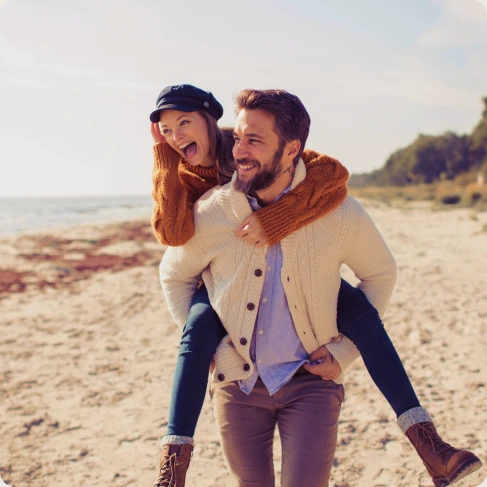 Ein Mann trägt eine Frau auf dem Rücken. Sie sind am Strand und beide lachen.